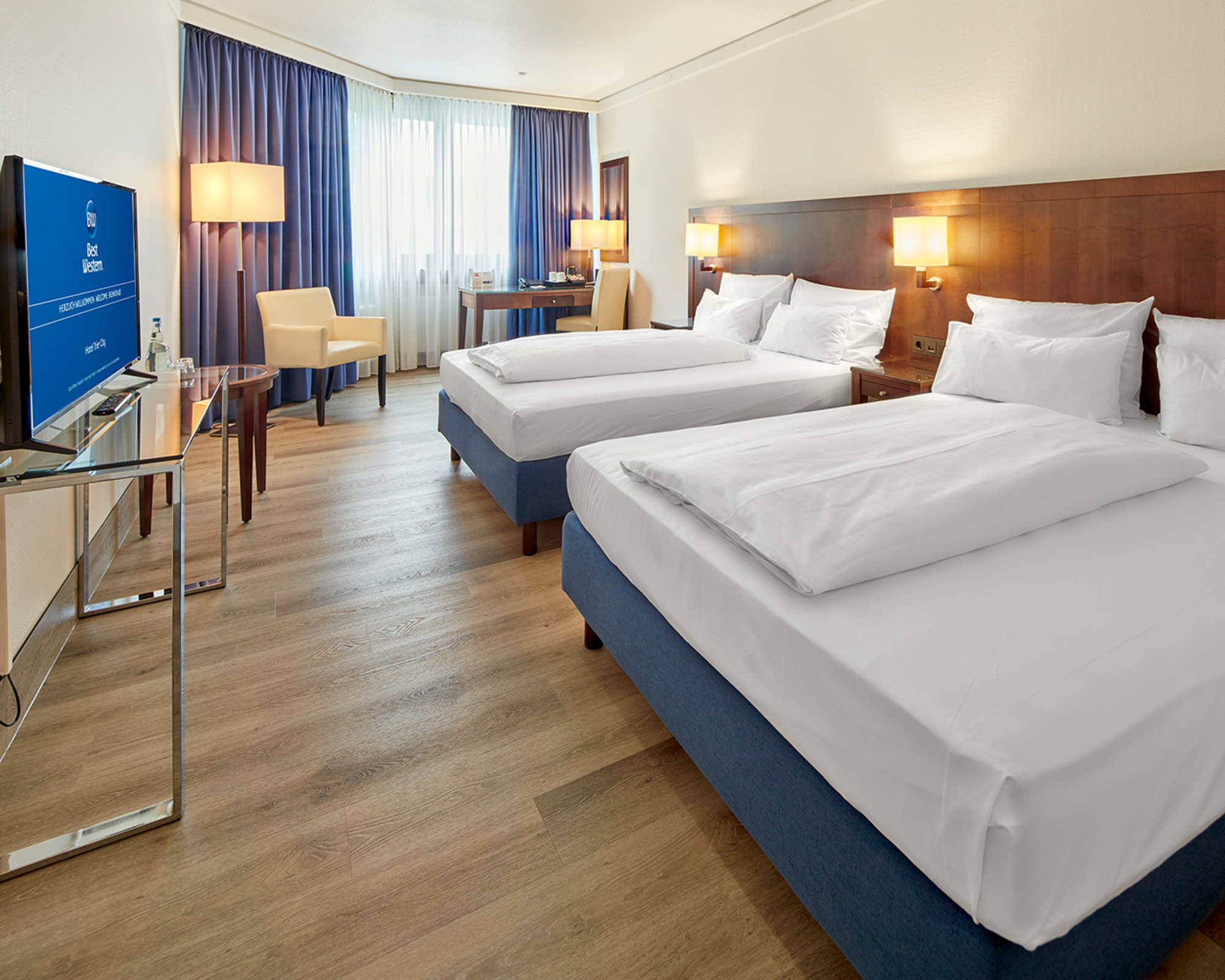 Lichtdurchlutetes Hotelzimmer mit zwei großen Betten, Sitzgelegenheit, TV und Schreibtisch